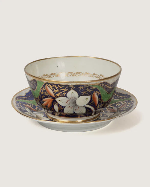 Front studio view of an antique British Imari style porcelain finger bowl sitting on its matching wide underplate, showing white flower blooms and gold detailing.